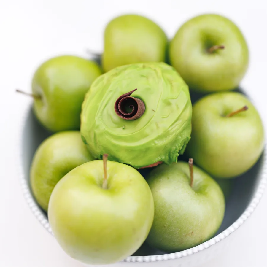 A green apple cake donut with glossy green apple icing nestled in a bowl of fresh green apples, from Michaels Bakery in Orrville, Ohio